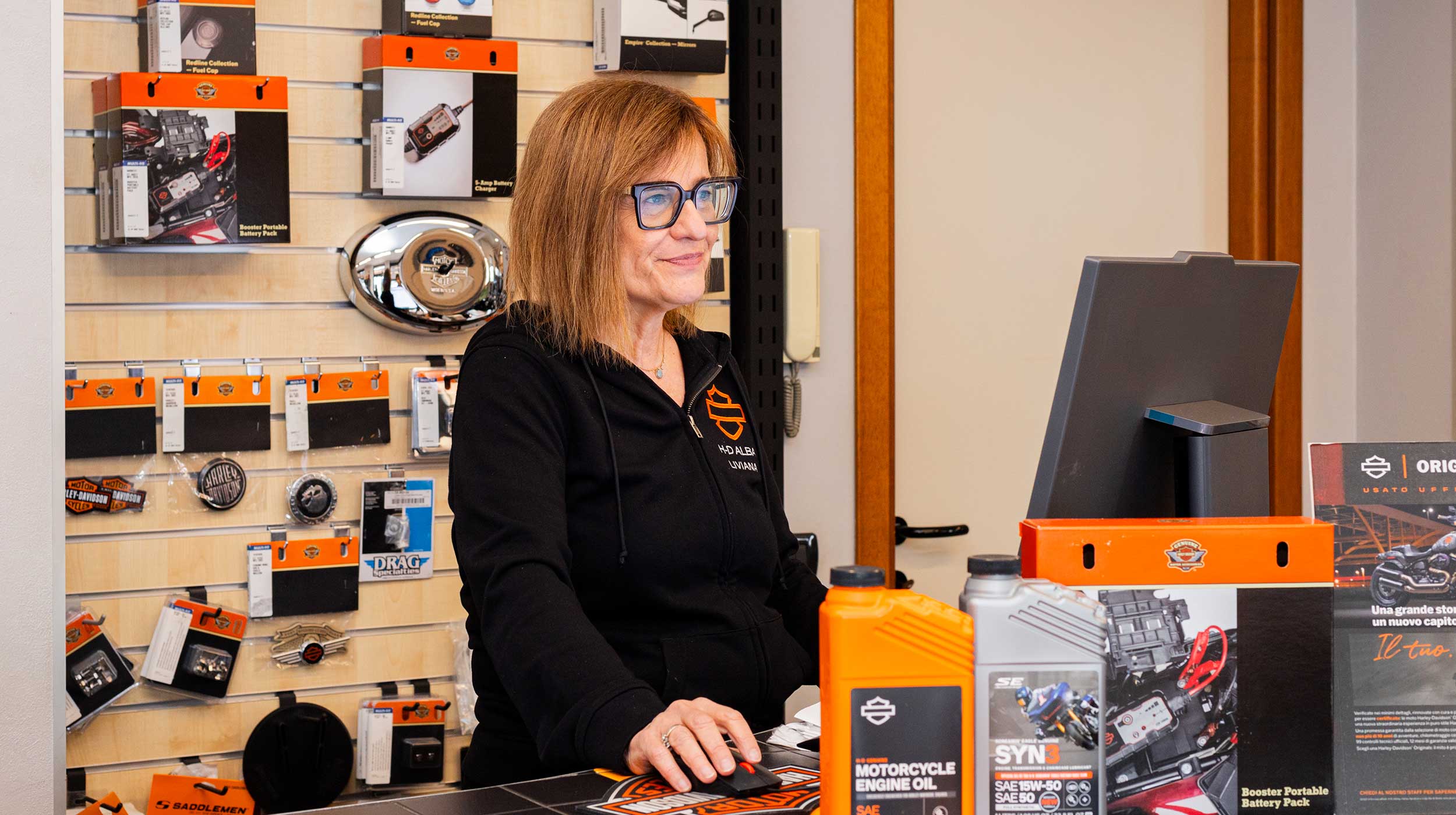 Servizio ric Addetta concessionaria Harley-Davidson Alba al banco ricambi e accessori, sorridente, con oli motore e prodotti originali in esposizione.